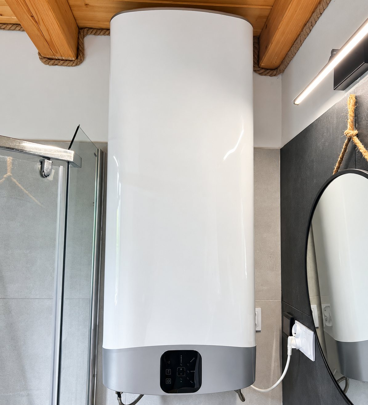 Un grand chauffe-eau électrique blanc et gris fixé au mur d'une salle de bain, près d'une douche vitrée et d'un miroir rond.
