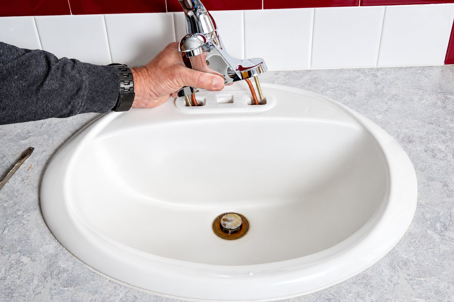 Une main tient un robinet mitigeur chromé au-dessus d'un lavabo blanc encastré dans un plan de travail gris, pendant son installation.
