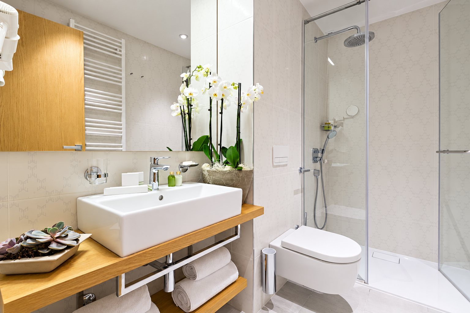 Salle de bain moderne et lumineuse avec une grande vasque blanche sur une tablette en bois, une douche vitrée, des serviettes roulées et des orchidées blanches.